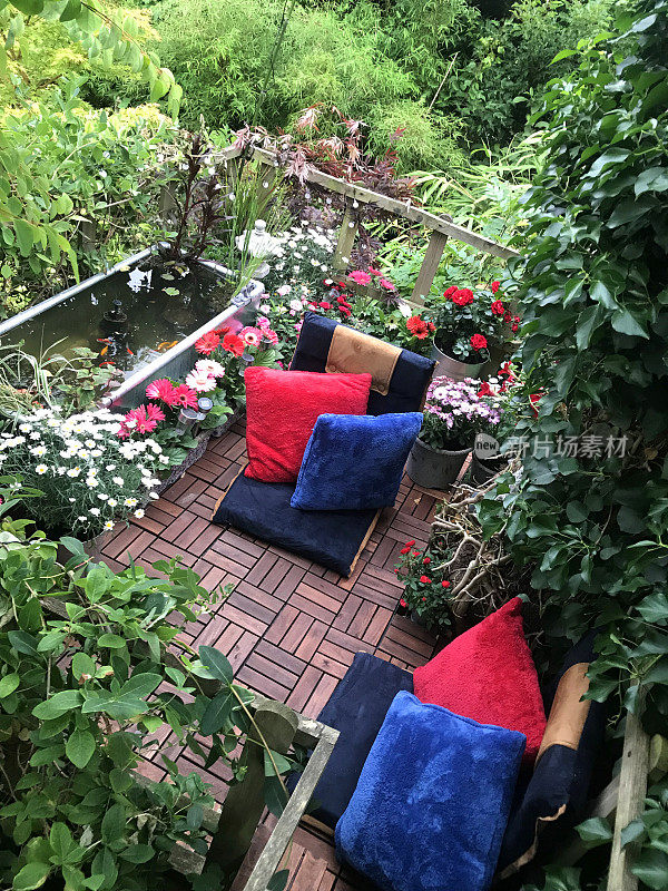 夏天的花园树屋阳台上有红色和蓝色的靠垫，锌池与太阳能喷泉泵，金鱼鱼，边缘植物，红色微型玫瑰，粉色非洲菊花，柚木装饰瓷砖，太阳能灯，照明