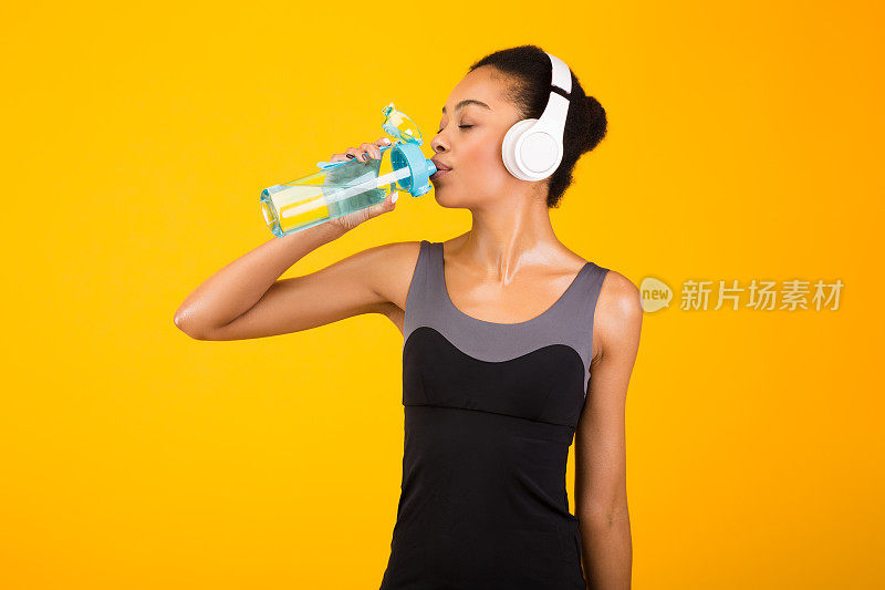 戴耳机的黑人年轻女士在黄色背景下喝水