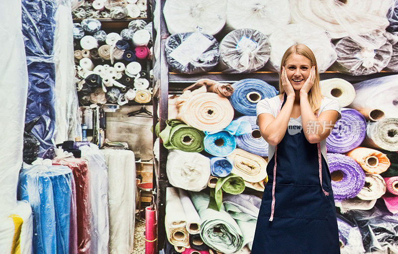 白人女手艺人穿着围裙，用卷尺站在服装店的布料店前