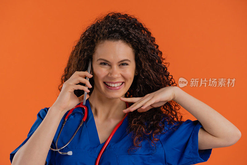 快乐年轻女医生看着摄像头，同时使用智能手机，橙色的背景