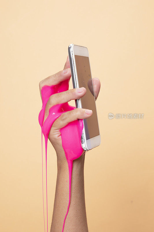 女人的手使用智能手机与霓虹粉黏泥玩具在橙色的流行艺术背景。