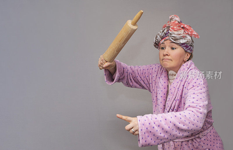 一名身穿睡袍、头戴围巾的女子手里拿着擀面杖，严肃地看着镜头。一个30-40岁的女人穿着家居服在一个灰色的背景，照片在工作室。