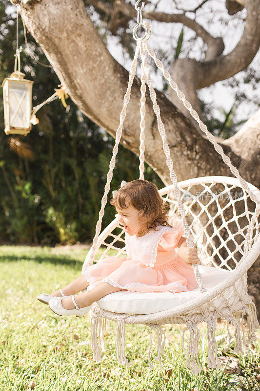 2019冠状病毒病期间，一名蹒跚学步的小女孩穿着一件复古的桃红色连衣裙，在家里度过她简单的2岁生日，快乐地坐在波西米亚树椅上