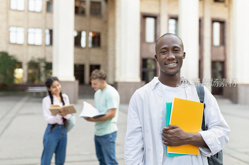 男非裔美国大学生背着背包和练习册在户外，在大学大楼附近摆姿势