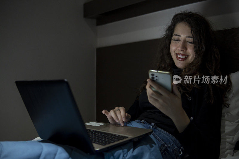 微笑的白人女孩在床上用电话，膝上放着笔记本电脑