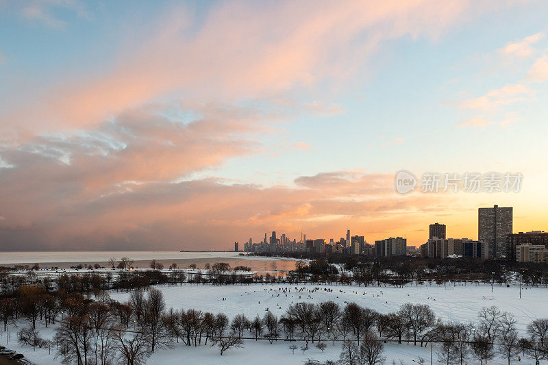 从蒙特罗斯海滩拍摄的芝加哥天际线的梦幻日落照片，前景中人们乘坐雪橇从蟋蟀山上滑下来，上面的蓝天中有粉红色和黄色的云。
