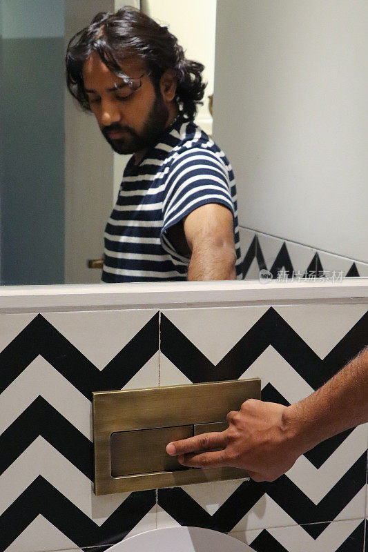 印度男子站在厕所前按下按钮冲水的镜子反射的特写图像，在黑色和白色之字形图案的墙砖上，集中在前景