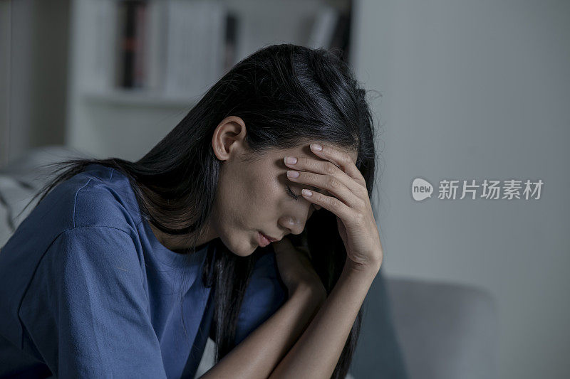 年轻的亚洲女性坐在家里的沙发上，感到悲伤、疲倦和担忧，心理健康状况堪忧。