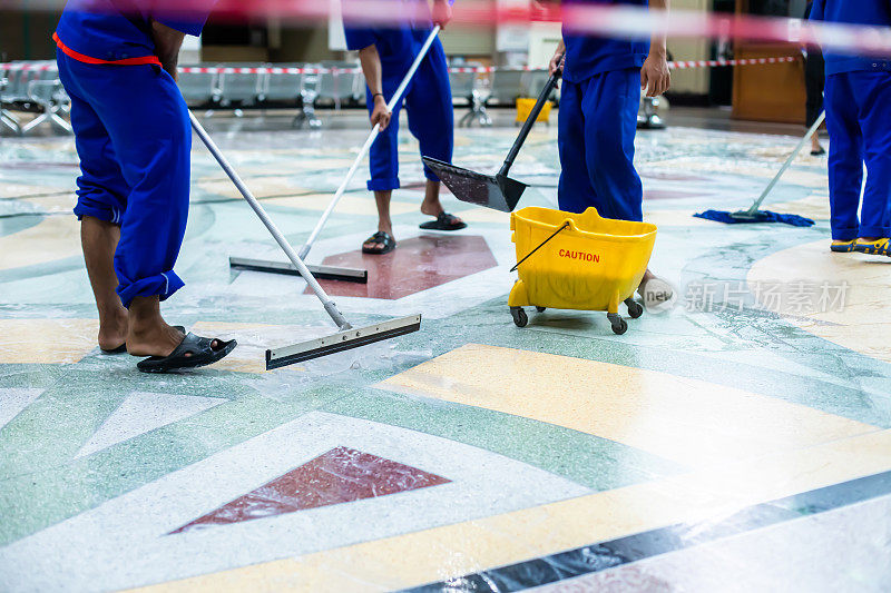 选择性地关注黄色桶的地板清洁剂和模糊的工作人员团队使用地板清洁剂，拖把和橡胶刮刀清洁。