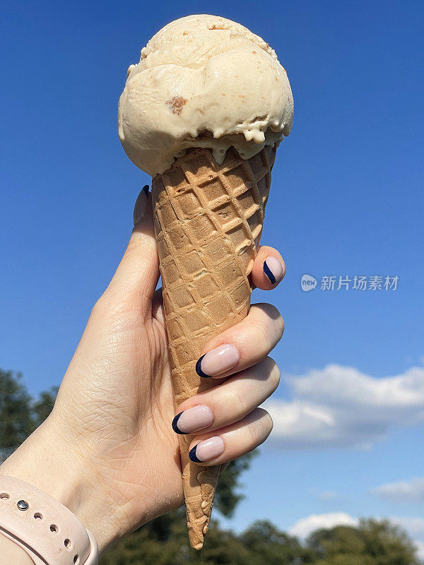 特写照片中，一个无法辨认的女人拿着华夫蛋筒，一勺太妃冰淇淋，黑色的指甲艺术，蓬松的云，阳光明媚，晴朗的蓝天，聚焦在前景上