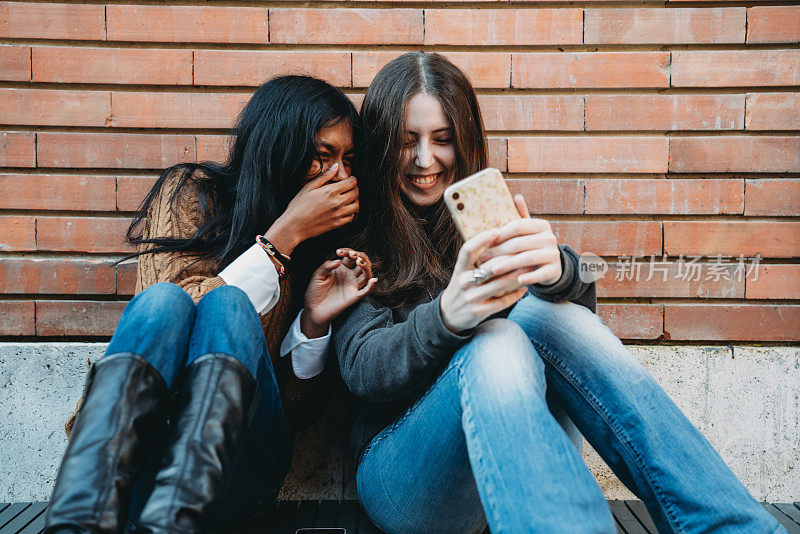 两个朋友靠砖墙坐在一起用手机
