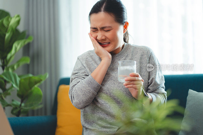 亚洲女性坐在客厅的沙发上，喝了凉水，摸了摸脸颊，就会感到牙齿敏感。