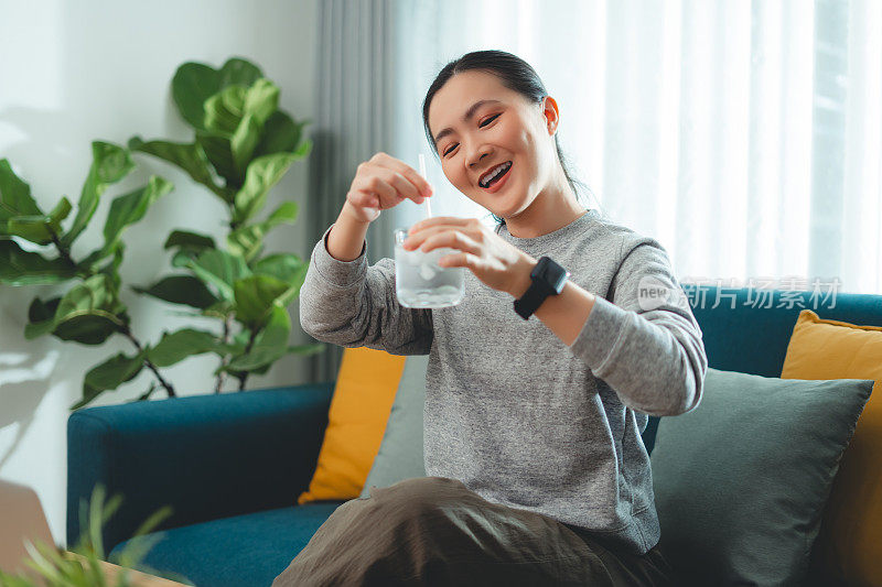 亚洲妇女坐在客厅的沙发上，用稻草搅拌水和冰休息。