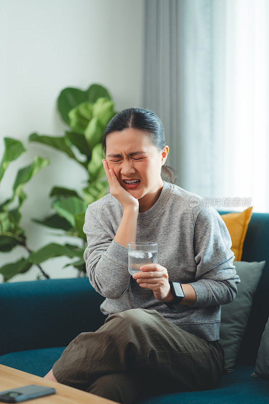 亚洲女性坐在客厅的沙发上，喝了凉水，摸了摸脸颊，就会感到牙齿敏感。