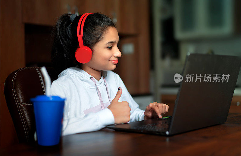 印度少女快乐的女孩戴着耳机，拿着笔记本电脑，在桌子上竖起大拇指