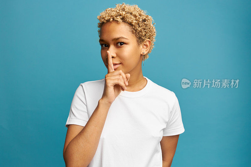 嘘!神秘的非洲年轻女子在蓝色的工作室背景上显示沉默的姿态，穿着白色t恤的拷贝空间为您的宣传内容，拥有金色短发