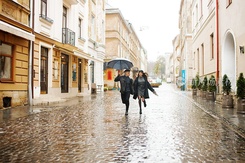 一对幸福的夫妇在雨天跑在街上。那个人手里拿着一把伞。2019年11月2日，乌克兰利沃夫。