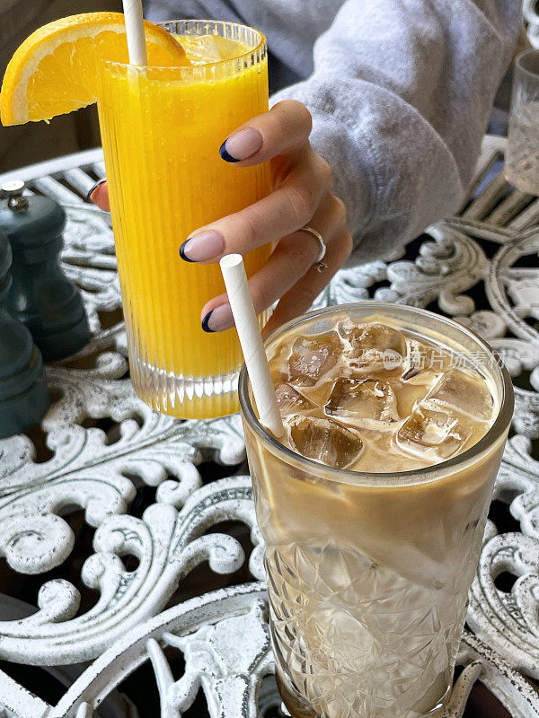 餐厅露天铁制桌子上的一杯冰咖啡饮料和吸管的特写图像，难以辨认的女人拿着一杯橙汁和橙片和纸吸管，重点放在前景