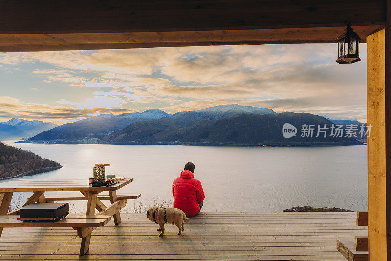 在挪威的日落时分，一名男子带着狗在露台上凝视着冬季峡湾的美景