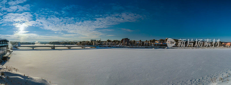 冰封的乌姆河和Lycksele当地城市景观的大桥全景，瑞典北部。冰封的河面上覆盖着刚下过的雪，冬日的阳光洒在蓝天上。