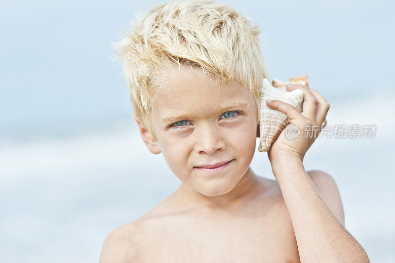 金发男孩在海滩上把贝壳举到耳边。