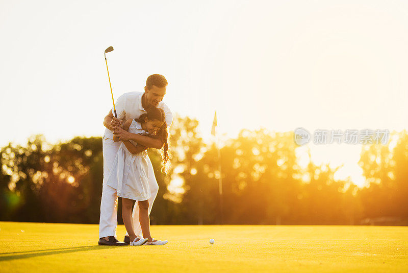 一个男人教他的女儿打高尔夫球。他引导着她，女孩正准备打高尔夫的第一拳