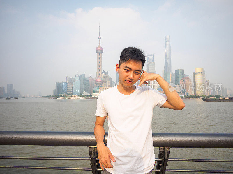 户外肖像的年轻亚洲人与上海外滩天际线的背景，使一个电话我的姿态。