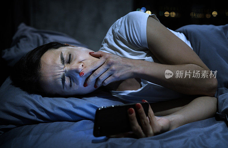 年轻困倦的女性使用手机，深夜打哈欠，躺在床上。智能手机成瘾的概念。