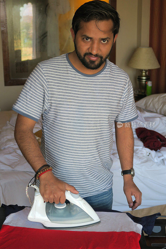 英俊的印度男子在酒店卧室的熨衣板上熨衣服，旁边是床和窗户，男子用电蒸汽熨烫t恤衣服，折叠，熨烫衣服，男子工作，洗家务，洗衣服戴rakhi手镯