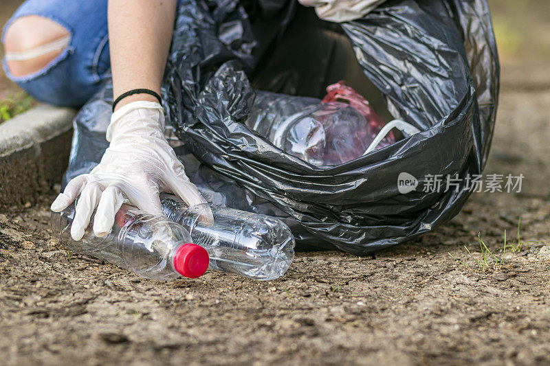 女志愿者穿着衣服在公园里捡垃圾和塑料垃圾。小女孩戴着手套，在户外把垃圾放进黑色的塑料袋里。