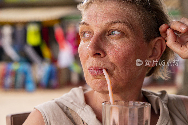 特写拍摄的欧洲50岁的女游客在沙滩上的露天餐厅，用吸管喝她的芒果鸡尾酒