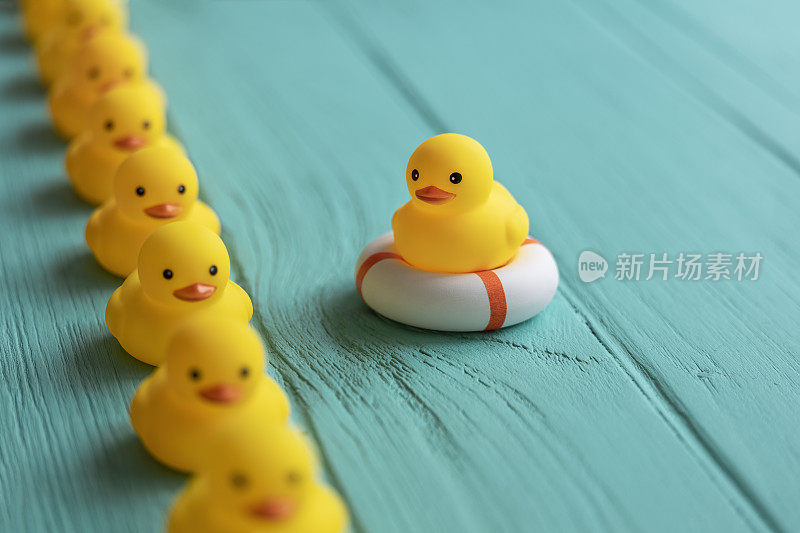 一排黄色的橡胶鸭子排成整齐的一排，一只鸭子坐在救生圈上，看着这一排鸭子经过，背景是风化的绿松石色的木质纹理，概念上代表着水。
