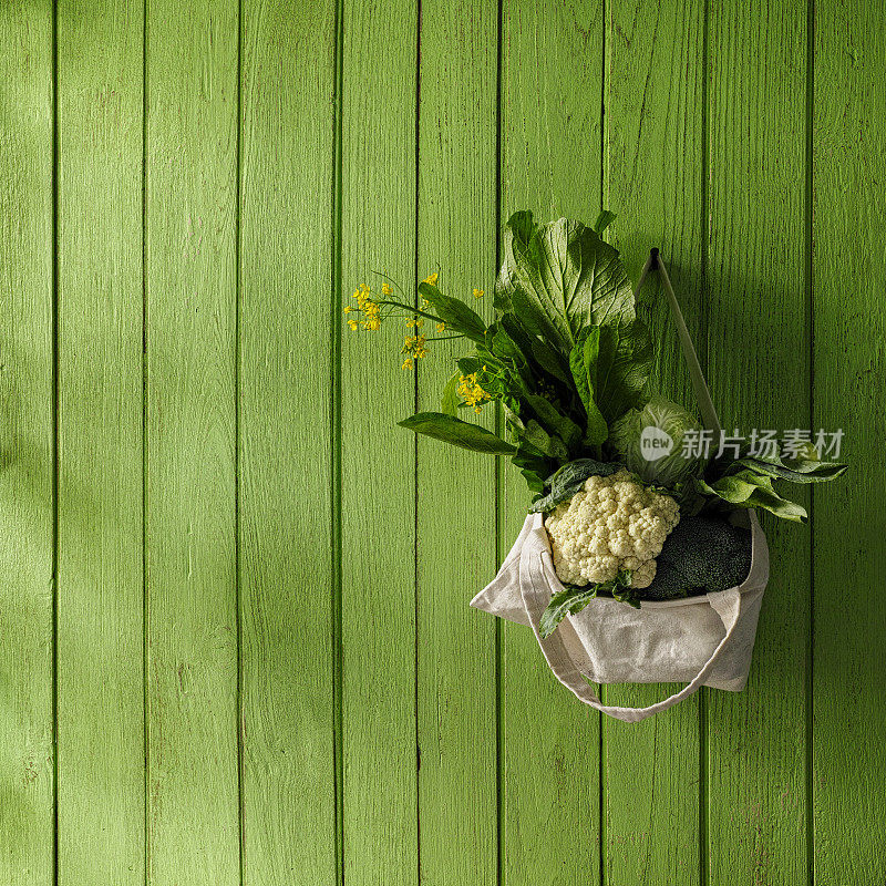 新鲜的绿叶蔬菜挂在一个可重复使用的棉花袋从钉子上，在一个旧的绿色木板墙的背景。