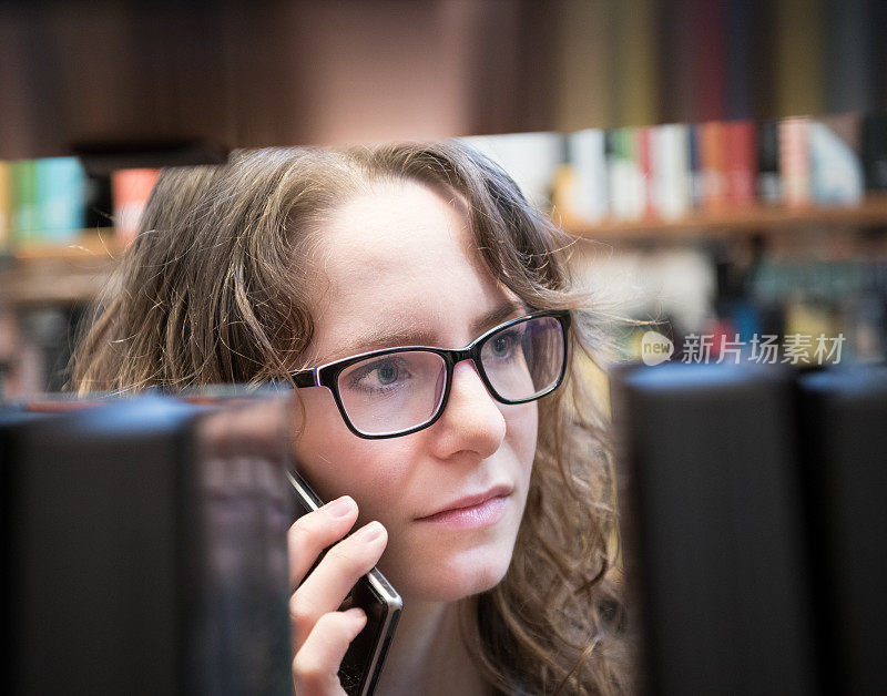 从书架上看，戴眼镜的学生在图书馆使用手机