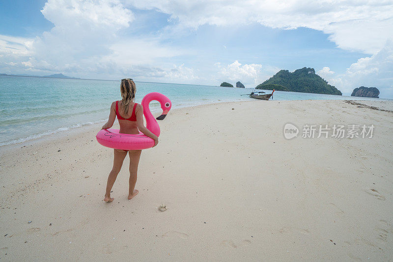 一名年轻女子与充气火烈鸟在泰国群岛田园诗般的海滩上散步。人们旅游目的地有趣和酷的态度概念