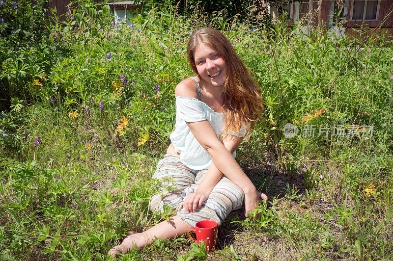 一个夏天的早晨，快乐的女孩坐在草地上，端着一杯红色的咖啡。