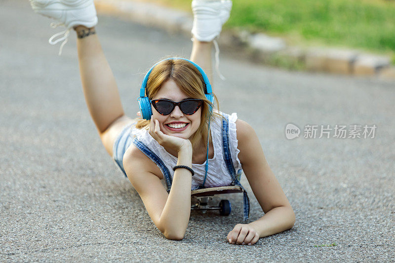 年轻女子戴着耳机坐在路上的滑板上。在一个阳光明媚的日子里，在公园里玩滑板的漂亮年轻女子。年轻迷人的美丽和时尚的年轻女人与滑板摆姿势的照片。她站在C里享受