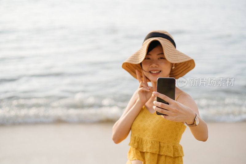 年轻的亚洲女游客戴着太阳帽用智能手机在海滩自拍