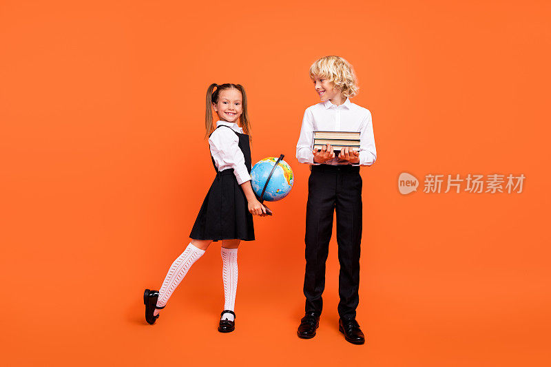 照片中的两个小学生拿着一堆书地球仪，准备上课，穿着校服，孤立的橙色背景
