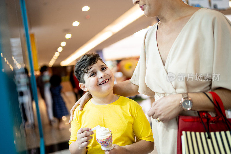 小男孩在吃冰淇淋，而他的妈妈在商场里看着橱窗