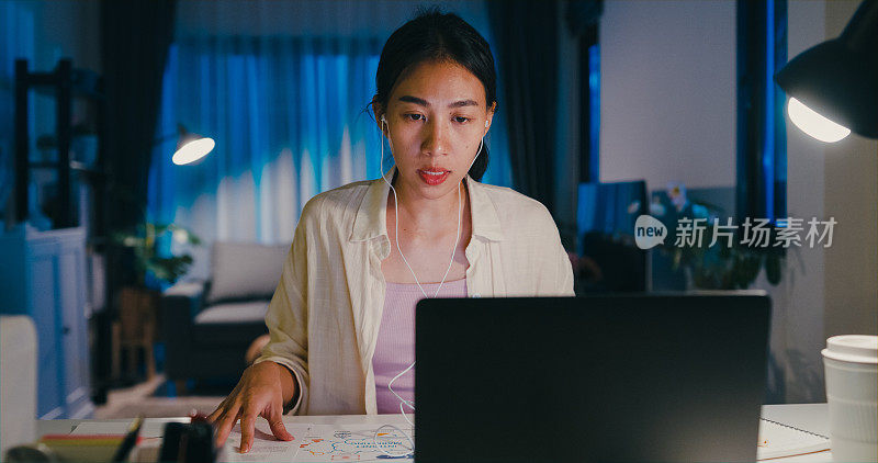 年轻的亚洲女性穿着正装衬衫和耳机坐在书桌前，用笔记本电脑写文件，在网上会议上做笔记，头脑风暴的想法，使报告加班到晚上在家。