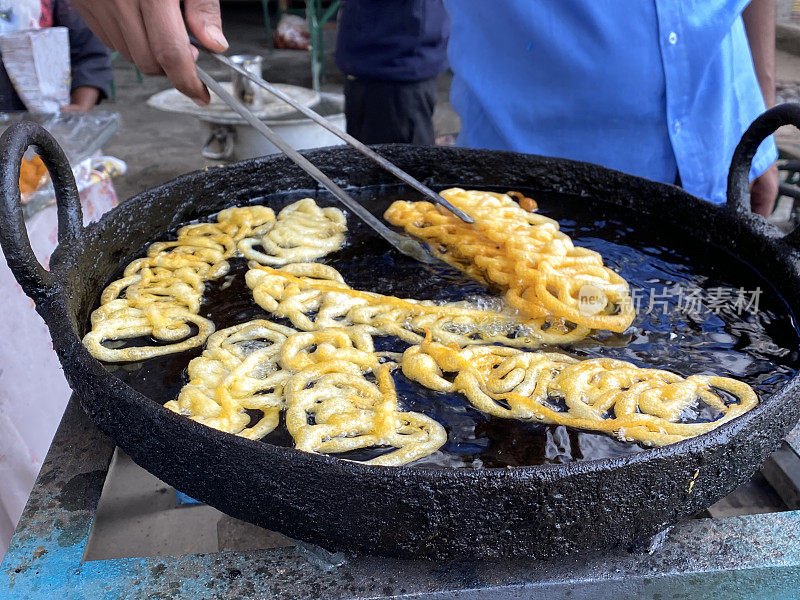 特写:贾勒比油炸面糊甜点，户外炉子上的街头小吃，厨师用钳子搅拌金黄色的螺旋面糊，煎锅里的油沸腾，高视图，聚焦前景