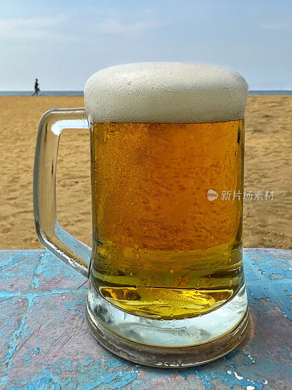 近距离图像的啤酒在玻璃酒杯覆盖的凝结与气泡形成泡沫的泡沫头在啤酒上，品脱玻璃在海滩酒吧户外桌子上，沙滩和清晰的蓝天背景，重点在前景
