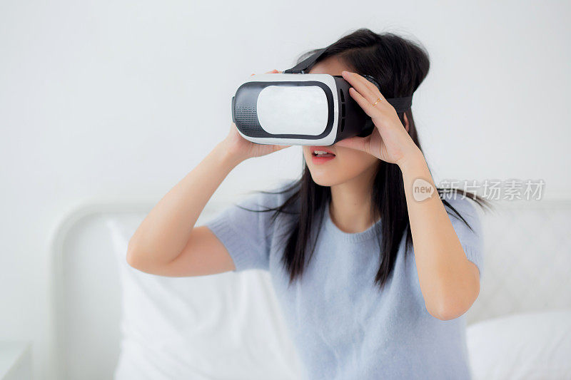 年轻的亚洲女性戴着vr耳机在家中卧室的床上兴奋地玩游戏，女性利用虚拟现实或虚拟世界创新模拟3D，生活方式和科技概念。