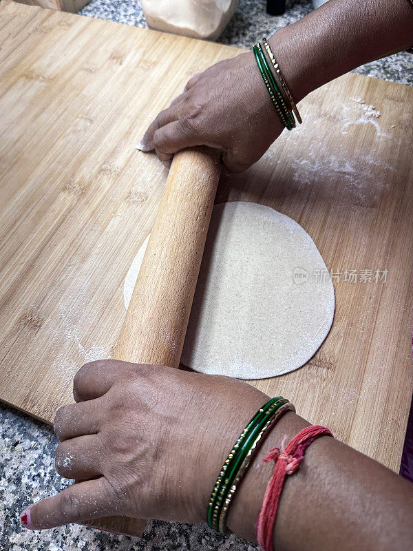 特写照片中，一名无法辨认的印度妇女用擀面杖在厨房柜台上的木制砧板上准备印度烤肉，将全麦atta面粉做成薄饼面团卷成圆盘，高架视角，聚焦于前景