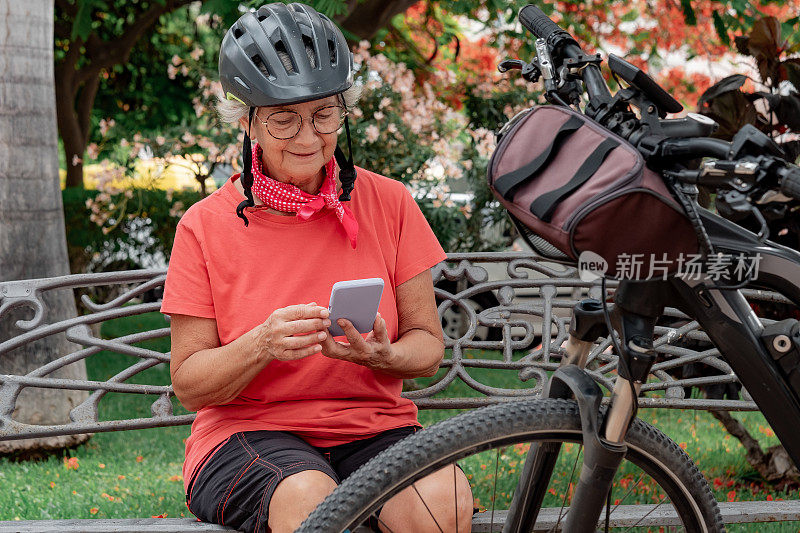 带着头盔微笑的骑自行车的老妇人坐在公园里，靠近她的电动自行车使用手机。享受健康生活方式的现代活跃祖母
