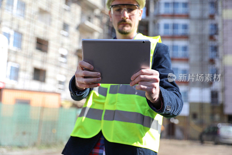 男建筑师在工地使用数码平板电脑，他穿着防护背心和头盔