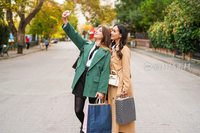 两名年轻女子手里拿着购物袋，在街上用手机自拍。购物的概念