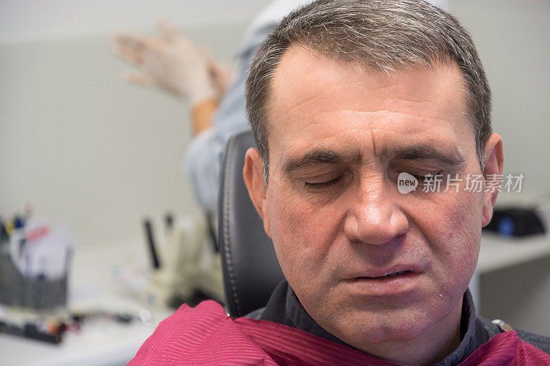 一个脸因疼痛而扭曲的男人在看牙医时闭上了眼睛。在模糊的背景中可以看到医生戴着医用手套的手。牙科治疗无痛的概念。