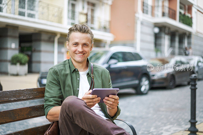技术融入日常生活，即人们利用数字设备进行信息和通信。金发男子在城市街道的长椅上用平板电脑搜索互联网信息。
